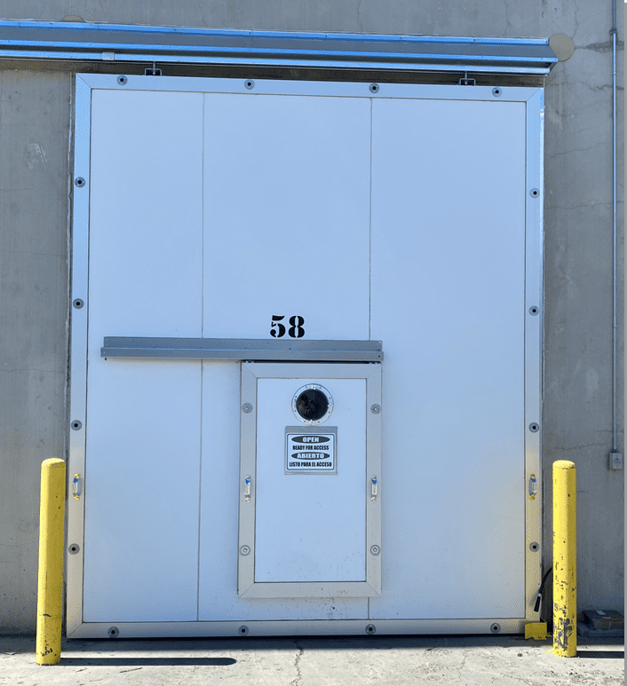 Large white industrial door with number 58, push bar, and a security porthole, flanked by yellow bollards